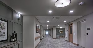 A modern, well-lit hallway in a senior living facility. The corridor features light gray walls, a patterned carpet, framed artwork, and a few light fixtures on the walls and ceiling. Doorways line both sides of the hall, leading to various rooms.