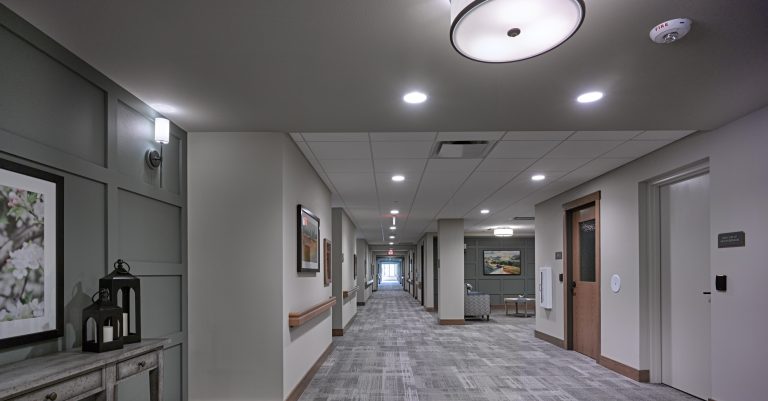 A modern, well-lit hallway in a senior living facility. The corridor features light gray walls, a patterned carpet, framed artwork, and a few light fixtures on the walls and ceiling. Doorways line both sides of the hall, leading to various rooms.