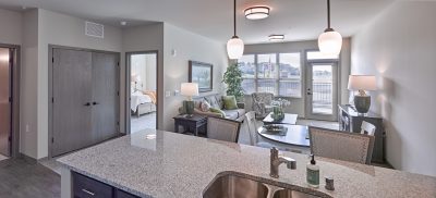 A modern, open-plan apartment living area with a kitchen island in the foreground. The living room features a sofa, chairs, a lamp, and a large window. A bedroom is visible through an open door. Light fixtures hang from the ceiling. Decor includes plants and a table lamp.