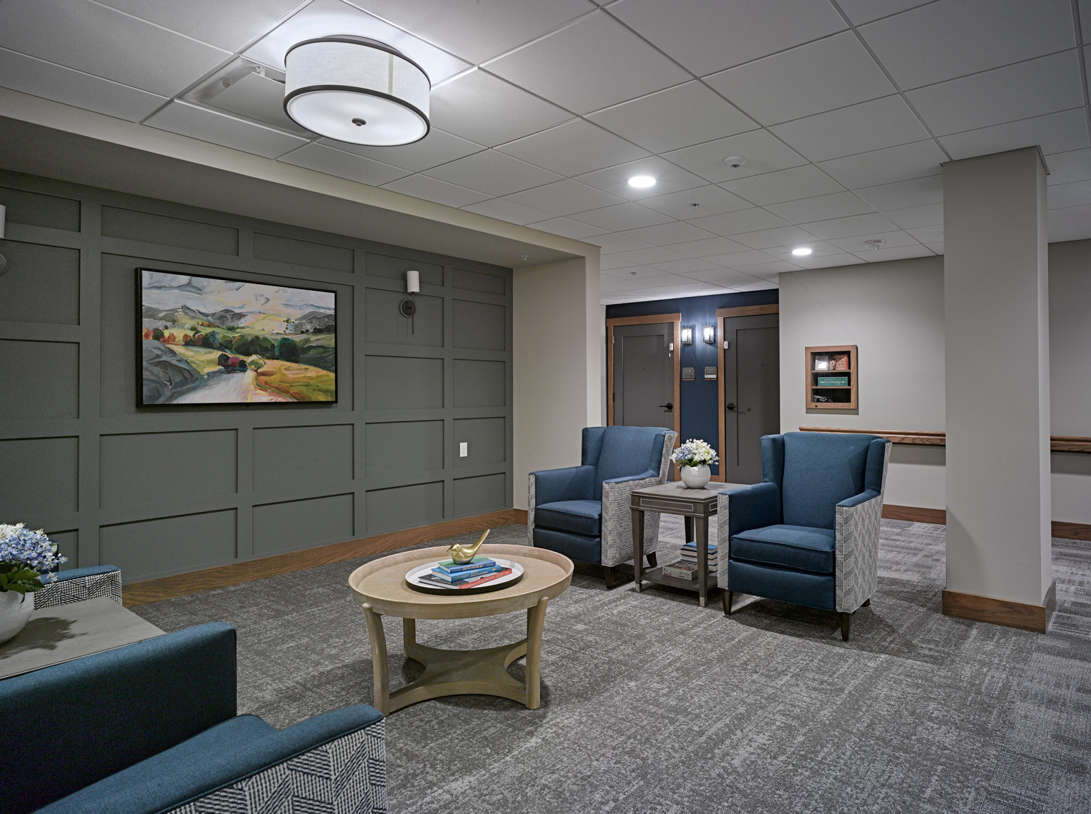 A well-lit, modern lounge area with grey walls, featuring panel molding and a landscape painting. The space includes blue and grey armchairs, a round wooden coffee table with books and a plant, grey carpeting, and a ceiling light fixture. Doorways are visible in the background.