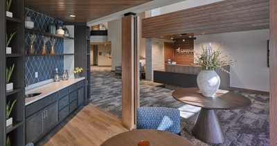 A modern senior living facility interior featuring a waiting area with round wooden tables and stylish chairs. The reception desk showcases a decorative vase with flowers. The décor includes shelves with various decorative items and a geometric tile wall pattern with dim lighting.