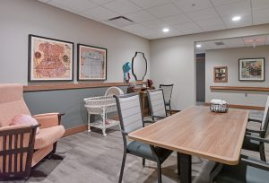 A well-lit room with light grey walls featuring a wooden table surrounded by five grey chairs. A pink armchair and a white wicker bassinet are in the corner. The walls display framed art, and there's a desk with a mirror and various items on it.