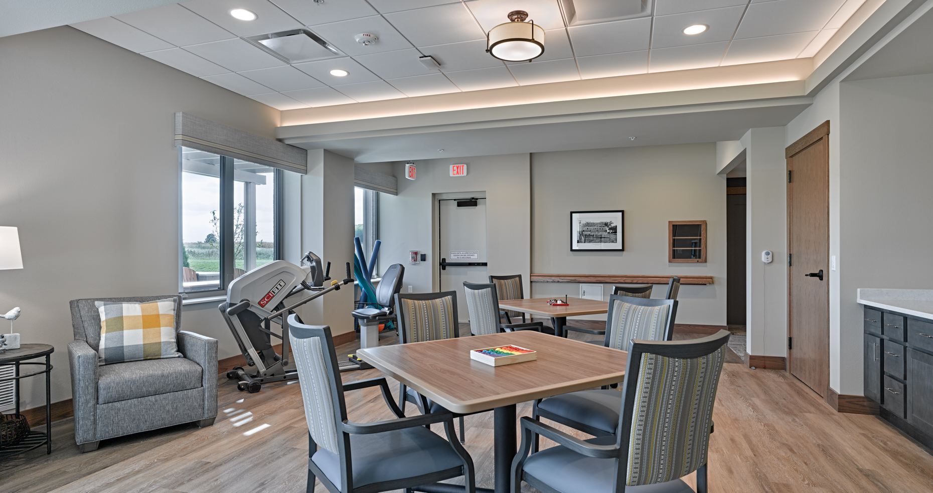 A modern, well-lit room with a workout machine, square tables, and chairs arranged for leisure. A single lounge chair with a plaid pillow is near the window. The walls have neutral shades, and framed pictures hang on the walls. A door leads out to a hallway.