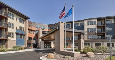 A modern senior living facility with multiple stories featuring a mix of light and dark colored siding. The entrance is covered with a wooden awning supported by brick columns. Two flagpoles stand nearby, displaying the American flag and a blue flag. Landscaping with rocks and plants surrounds the area.