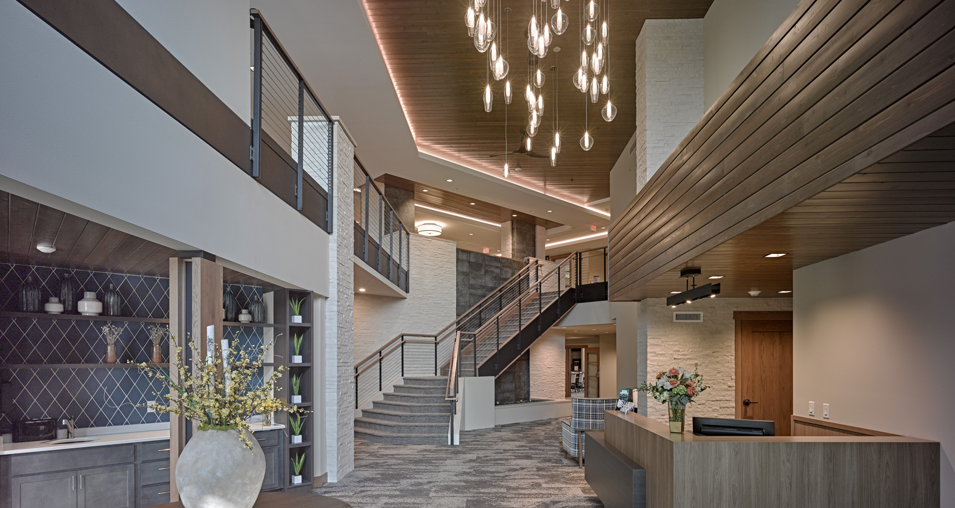 Modern senior living lobby with a stylish chandelier, contemporary furniture, and a spiral staircase leading to the upper floor. The decor includes wooden accents, a reception desk with flowers, and shelving displaying decorative items. The lighting creates a welcoming ambiance.