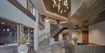 Modern senior living lobby with a stylish chandelier, contemporary furniture, and a spiral staircase leading to the upper floor. The decor includes wooden accents, a reception desk with flowers, and shelving displaying decorative items.