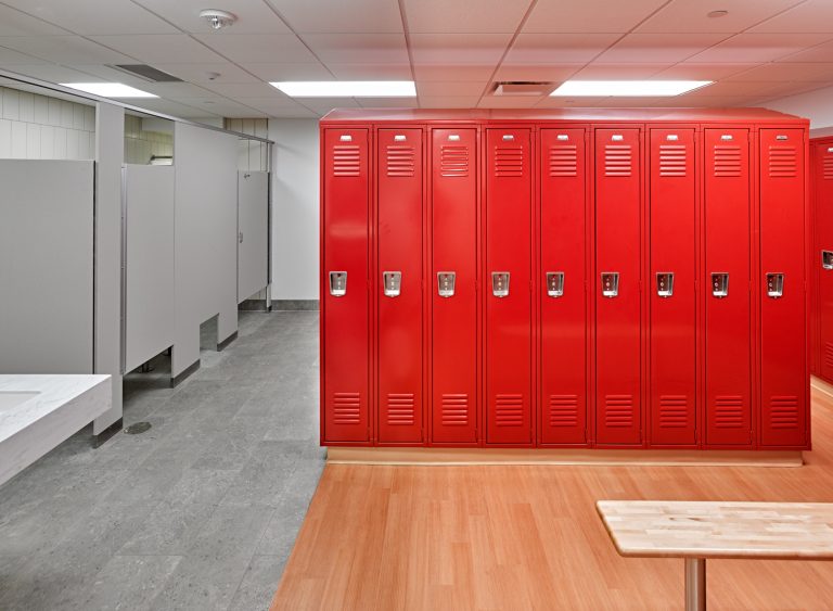 A clean locker room with a row of red lockers on a light wooden floor. To the left, there are gray bathroom stalls and a white sink. The ceiling is lined with white panels and recessed lighting, giving the room a bright and modern appearance.