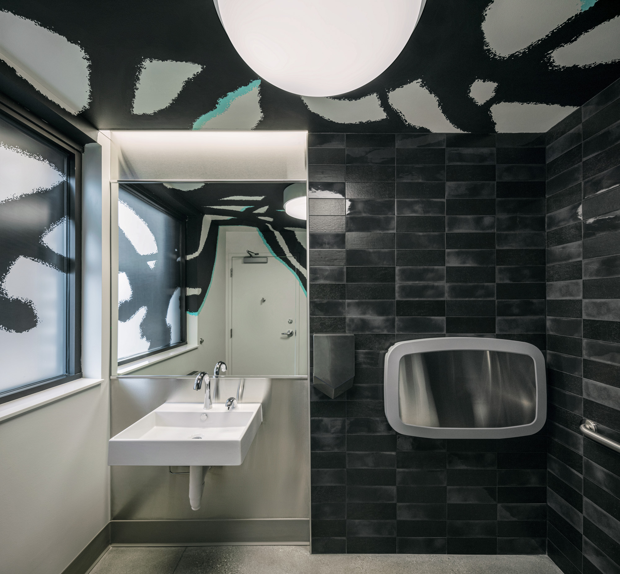 Restroom with black tiles wall, white and black patterned ceiling, a stainless steel baby changing table and concrete floors