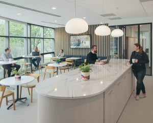 A modern, open office space with large windows and natural light. Four people are sitting at round tables and one is standing at a large, marble-topped island with hanging lights. There are plants on the tables and island. A large screen is on the back wall.