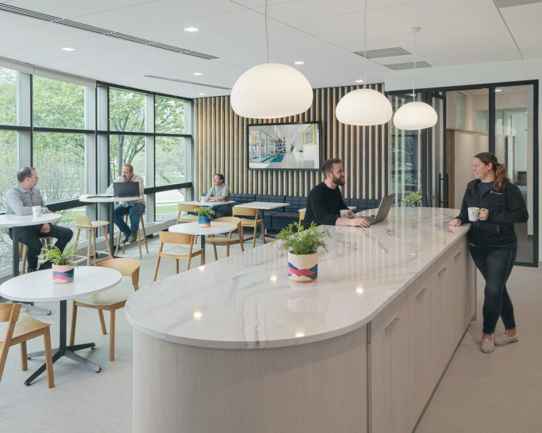 A modern, open office space with large windows and natural light. Four people are sitting at round tables and one is standing at a large, marble-topped island with hanging lights. There are plants on the tables and island. A large screen is on the back wall.