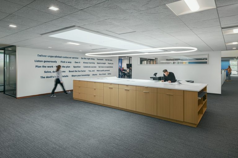 A modern office space with a large central desk and an employee working on it. Another person is walking past the desk. The wall behind features motivational statements in blue font. The ceiling has recessed lighting, and the area is spacious with grey flooring.