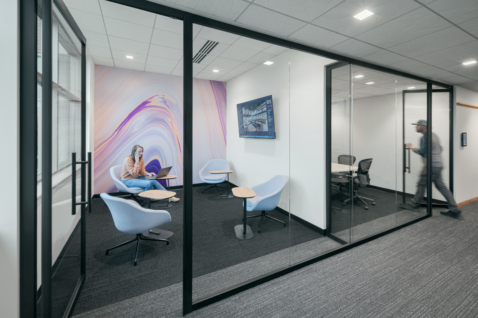 Modern office space with glass walls, showcasing a small lounge area and a separate meeting room. A person sits in the lounge on a blue chair, working on a laptop. Another person is walking down the hallway. Abstract artwork and a flat-screen TV adorn the walls.