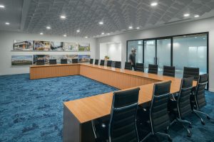 A modern conference room featuring a large U-shaped wooden table surrounded by black ergonomic chairs on a blue carpet. The back wall displays architectural images. A person is faintly visible walking past the glass doors in the background.