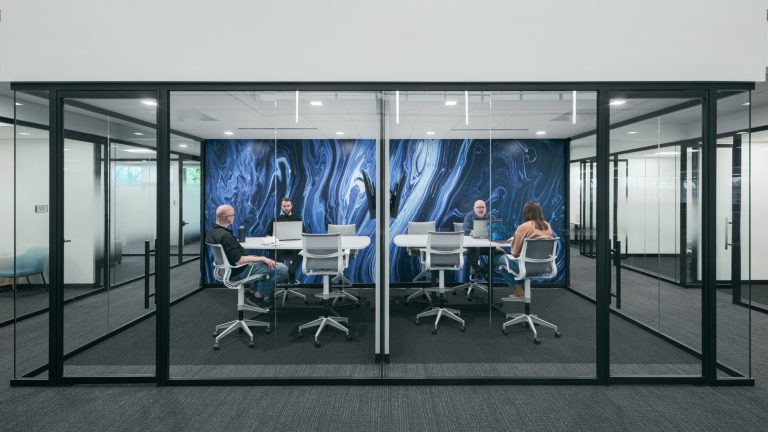 Four individuals are seated around a table in a modern conference room enclosed with glass walls. The room features a large, abstract blue and black mural on one wall. Each person is focused on their laptops or engaged in discussion.