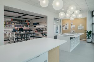 A modern office space featuring two people sitting at a table conversing in a library-like area with extensive shelving holding books and materials. The foreground has a white table and shelving unit, and the background includes a bulletin board and indoor plants.