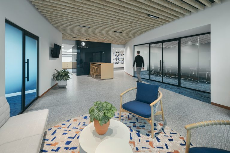 A modern office lobby featuring a small seating area with blue chairs and a white sofa around a round table with a plant. A person is walking past, and there are glass-walled meeting rooms on the right. The ceiling has exposed wooden beams.