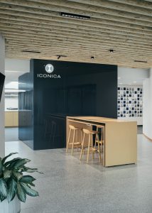 Modern office space featuring a wooden high table with stools, a black glass wall with the "ICONICA" logo, and a ceiling with exposed wooden beams. A potted plant is in the foreground, and a wall displaying numerous small photos is in the background.