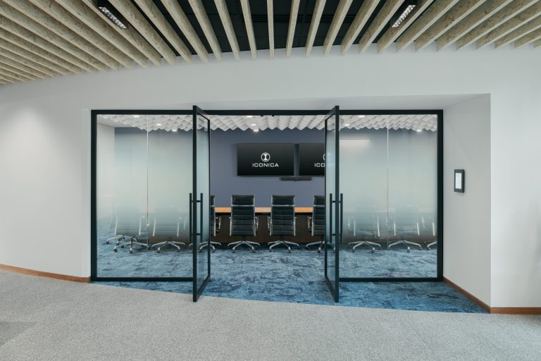 A modern conference room with glass doors open, revealing a long wooden table surrounded by black office chairs. The room has blue carpet and a TV on the back wall displaying the logo "ICONICA." The ceiling features a stylish grid design.