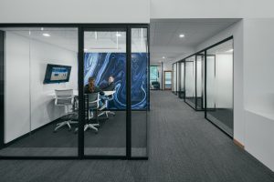 A modern office hallway with carpeted flooring and glass-walled meeting rooms. One meeting room is visible, occupied by three people seated around a table. A large screen on the wall shows a presentation, and a blue and black swirling mural serves as a backdrop.