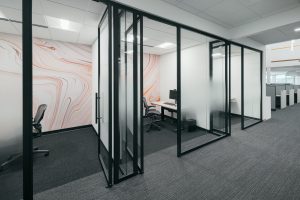 An office hallway with glass-walled private rooms on the left, featuring modern minimalist chairs and desks. The walls inside the rooms are decorated with a white and orange marble pattern. Cubicles are visible in the background to the right.