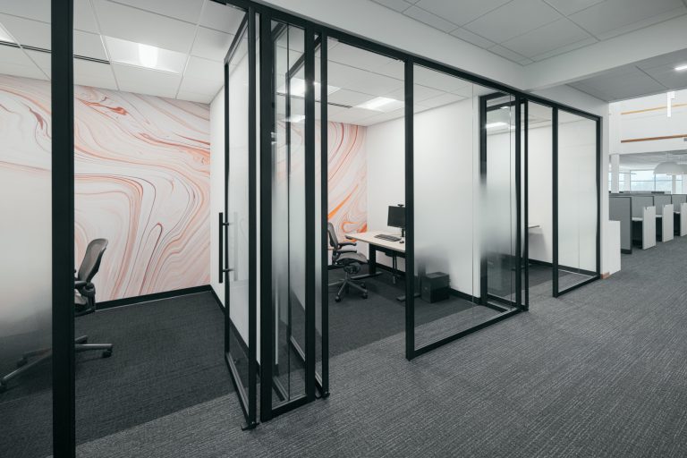 An office hallway with glass-walled private rooms on the left, featuring modern minimalist chairs and desks. The walls inside the rooms are decorated with a white and orange marble pattern. Cubicles are visible in the background to the right.