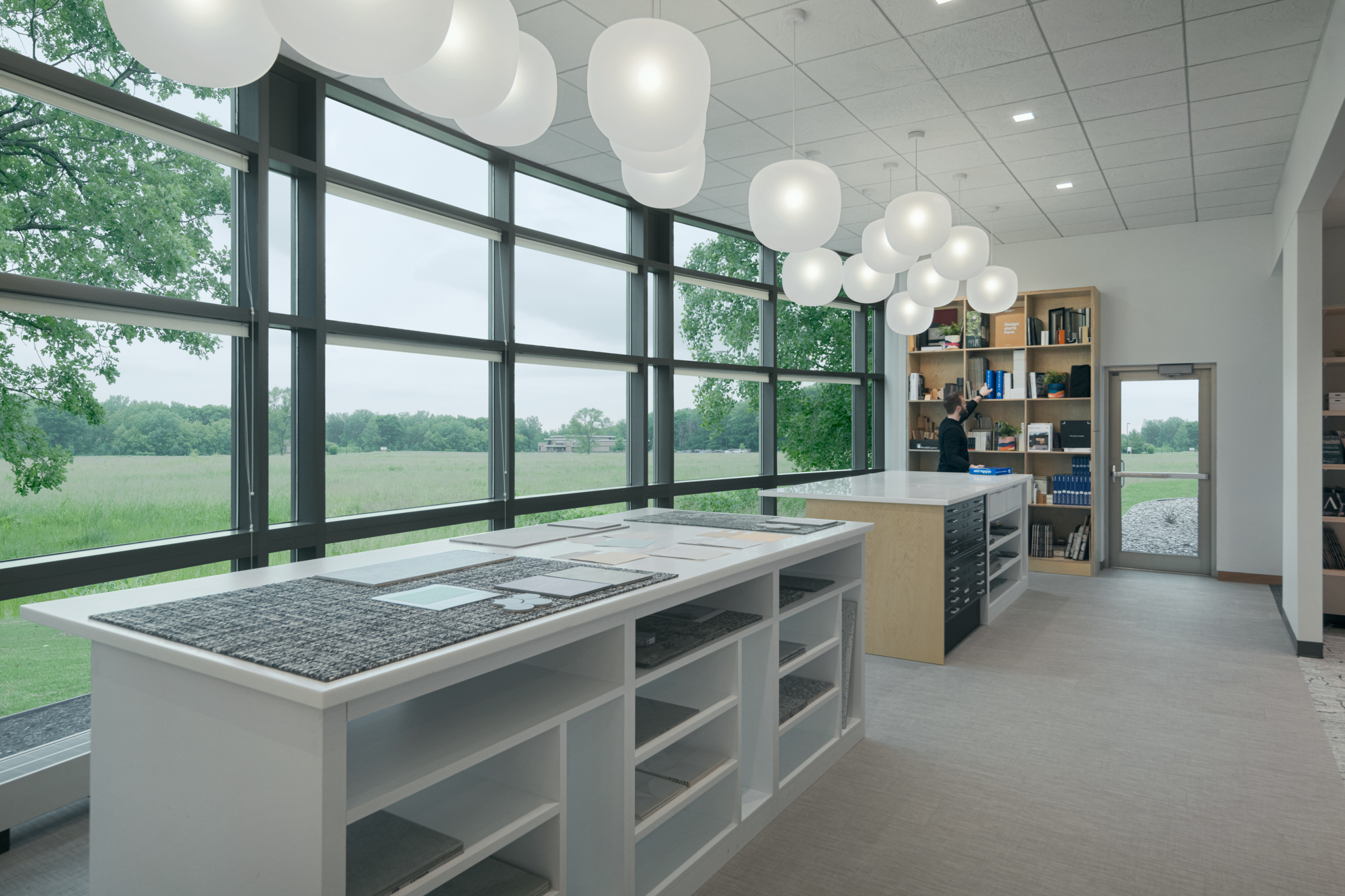 A modern library or office space with large windows offering a view of a green landscape. The room features several bookshelves, tables with storage, and hanging white spherical lights. A person is browsing through books on a shelf in the background.