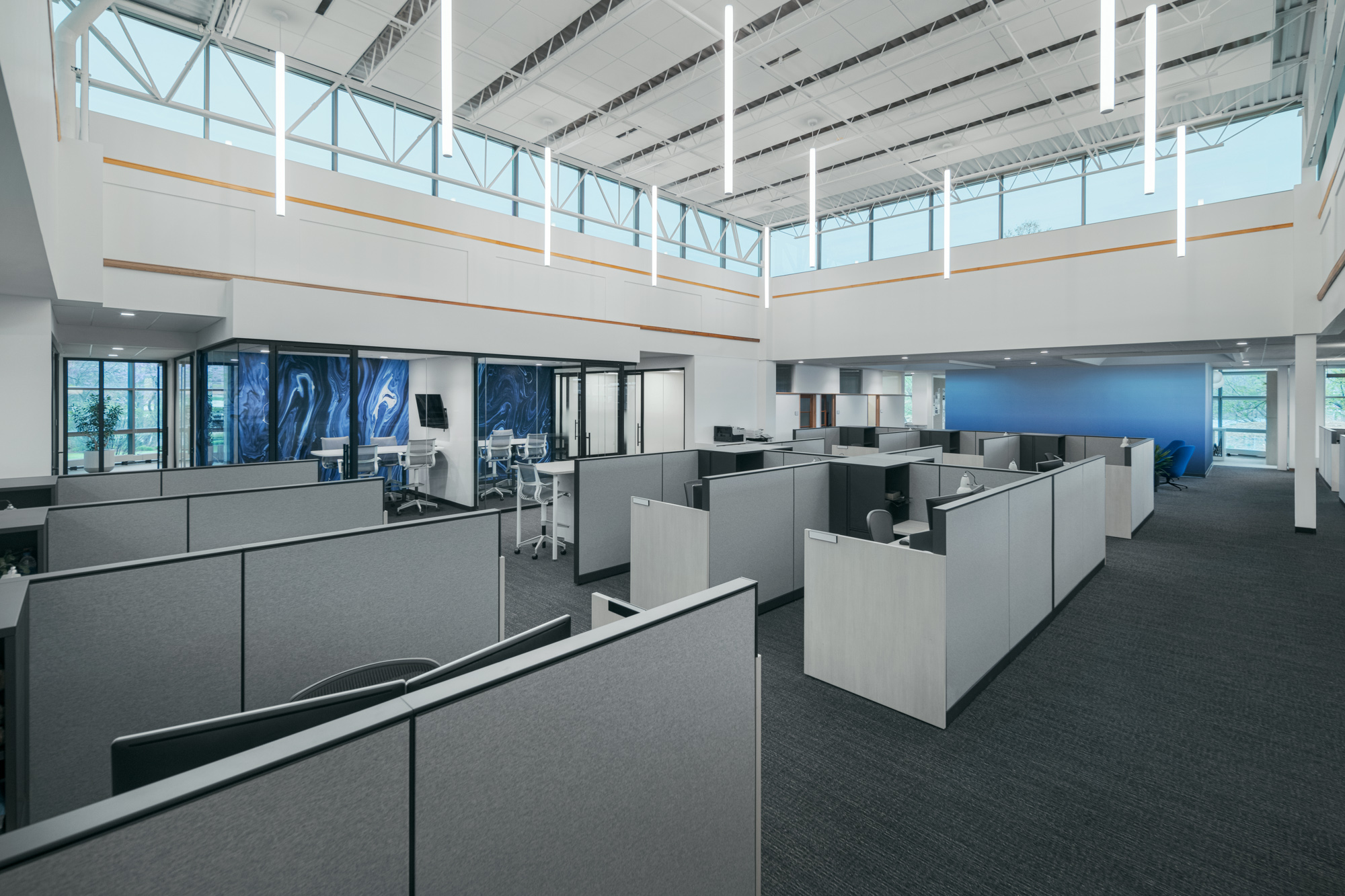 A spacious, modern office with high ceilings and large windows. The office features an open floor plan with gray cubicles and workstations. Natural light fills the area, which includes several standing desks and ergonomic chairs. Vertical lights hang from the ceiling.