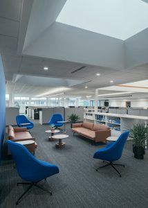 A modern office lounge area with brown leather couches and blue chairs arranged around small, round coffee tables. Potted plants are placed beside the arrangement. Behind the seating area, open-plan office cubicles are visible under a daylight ceiling feature.