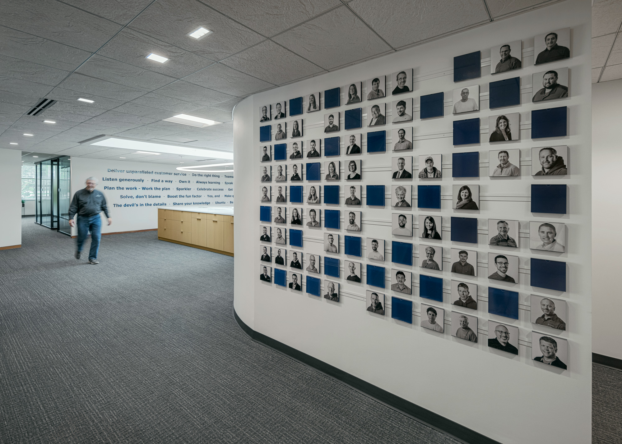 A hallway with gray carpeting and white walls features a display of black and white employee portraits arranged in a grid. A person walks by in the background, and there are motivational quotes on the far wall.