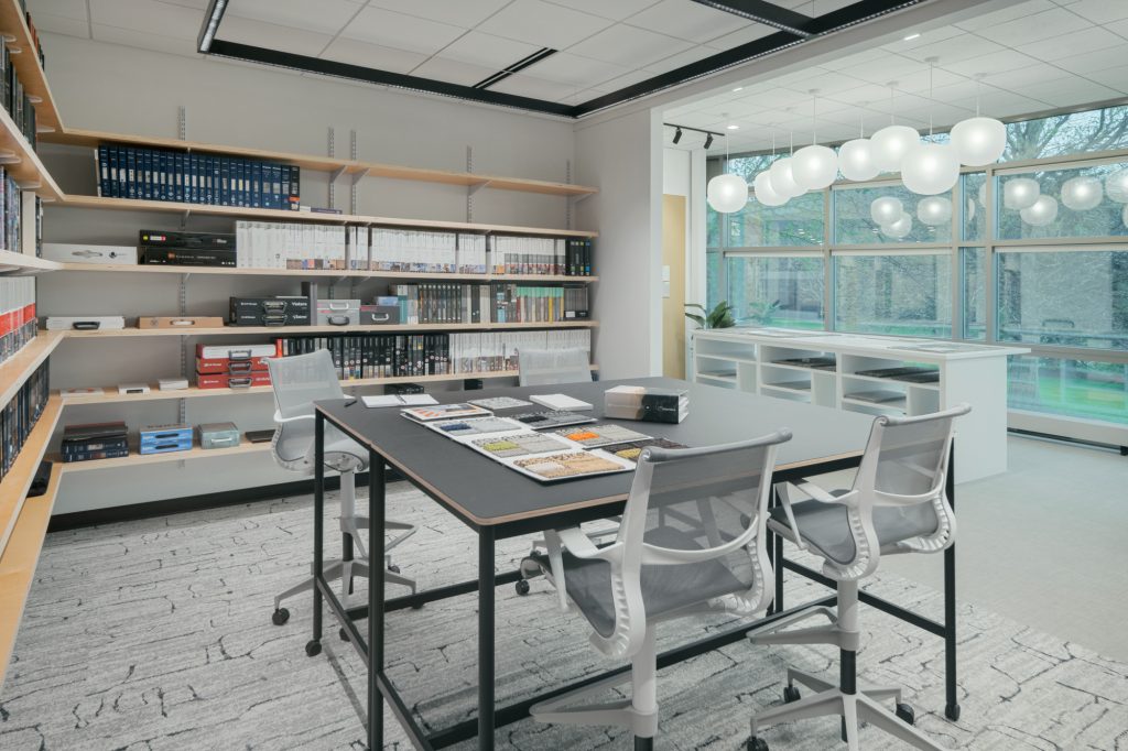 A modern design library with large windows, overhead spherical lights, and wall-mounted bookshelves filled with books and magazines. A black table surrounded by four gray chairs holds various open books and materials. Light grey carpeting covers the floor.