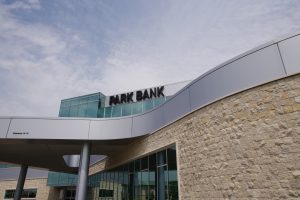 The exterior of a modern building with a stone and glass facade under a partly cloudy sky. A sign on the building reads "Park Bank." The structure features large windows and a curved architectural design.