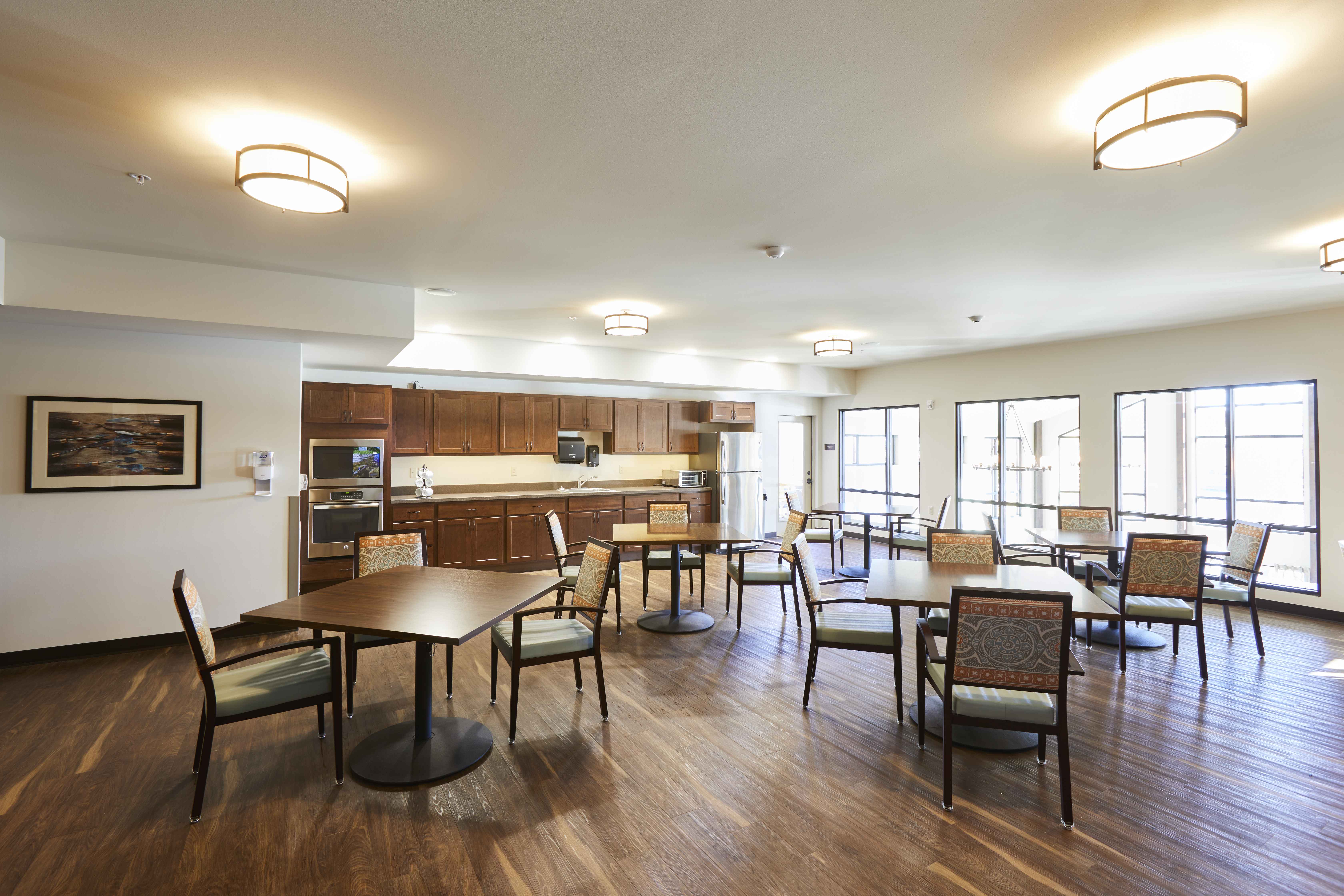 A spacious room with wooden flooring, warm lighting, and several wooden tables and chairs. The far end has a kitchen area with cabinets, a sink, and appliances. Large windows let in natural light. Artwork and a water dispenser are on the left wall.