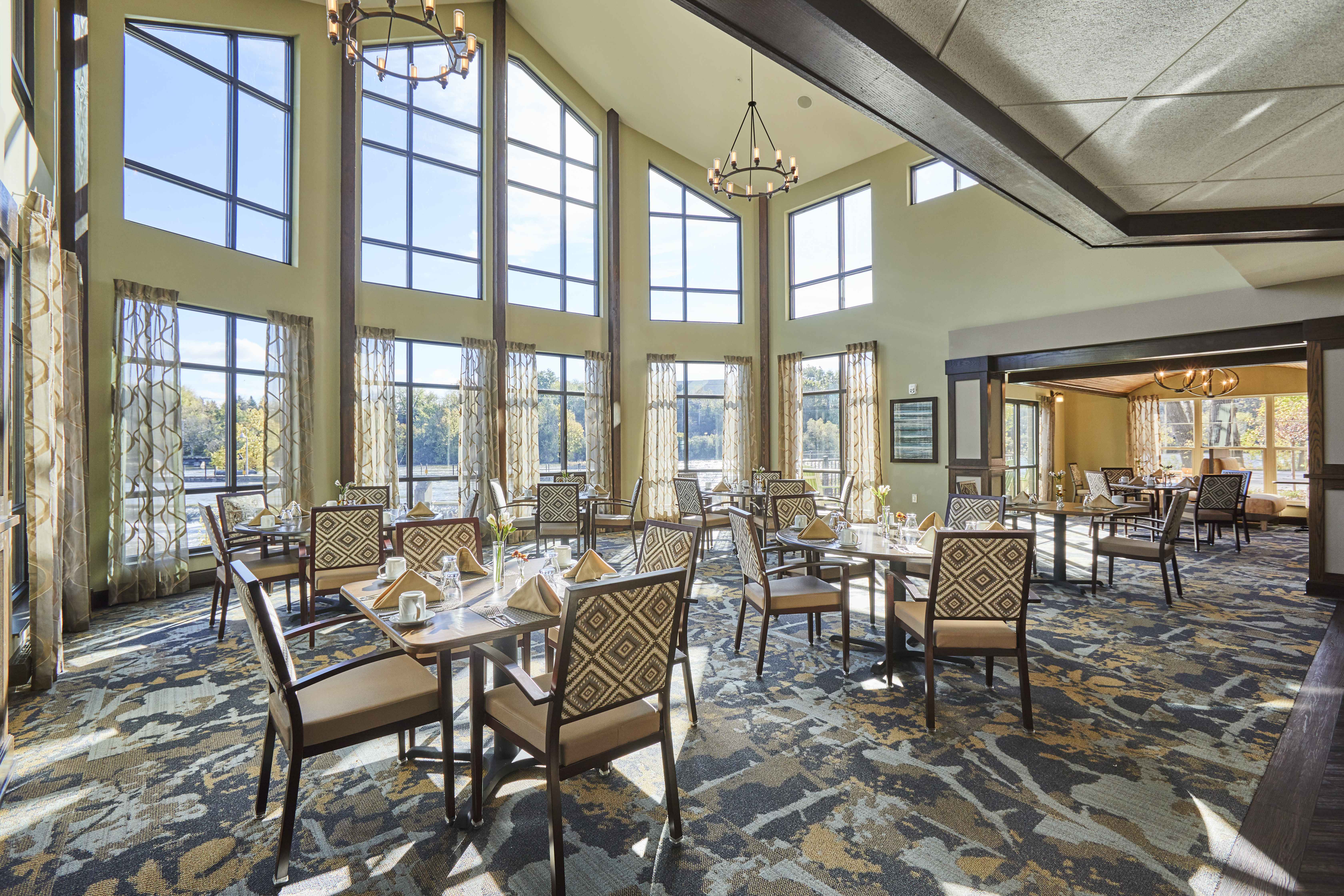 Senior living facility dining room with chandeliers, patterned carpet, and expansive floor to ceiling windows with a view of the river