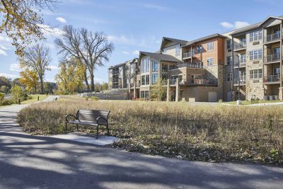 A senior living facility on a sunny fall day with a walking path, park bench and expansive windows with a view of the lake