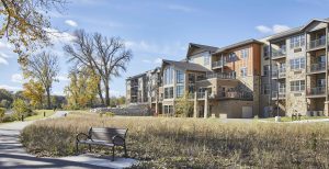 A senior living facility on a sunny fall day with a walking path, park bench and expansive windows with a view of the lake