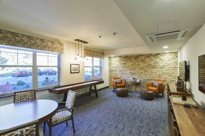 Senior living facility game room with a brick accent wall, blue gray carpet, and shuffle board