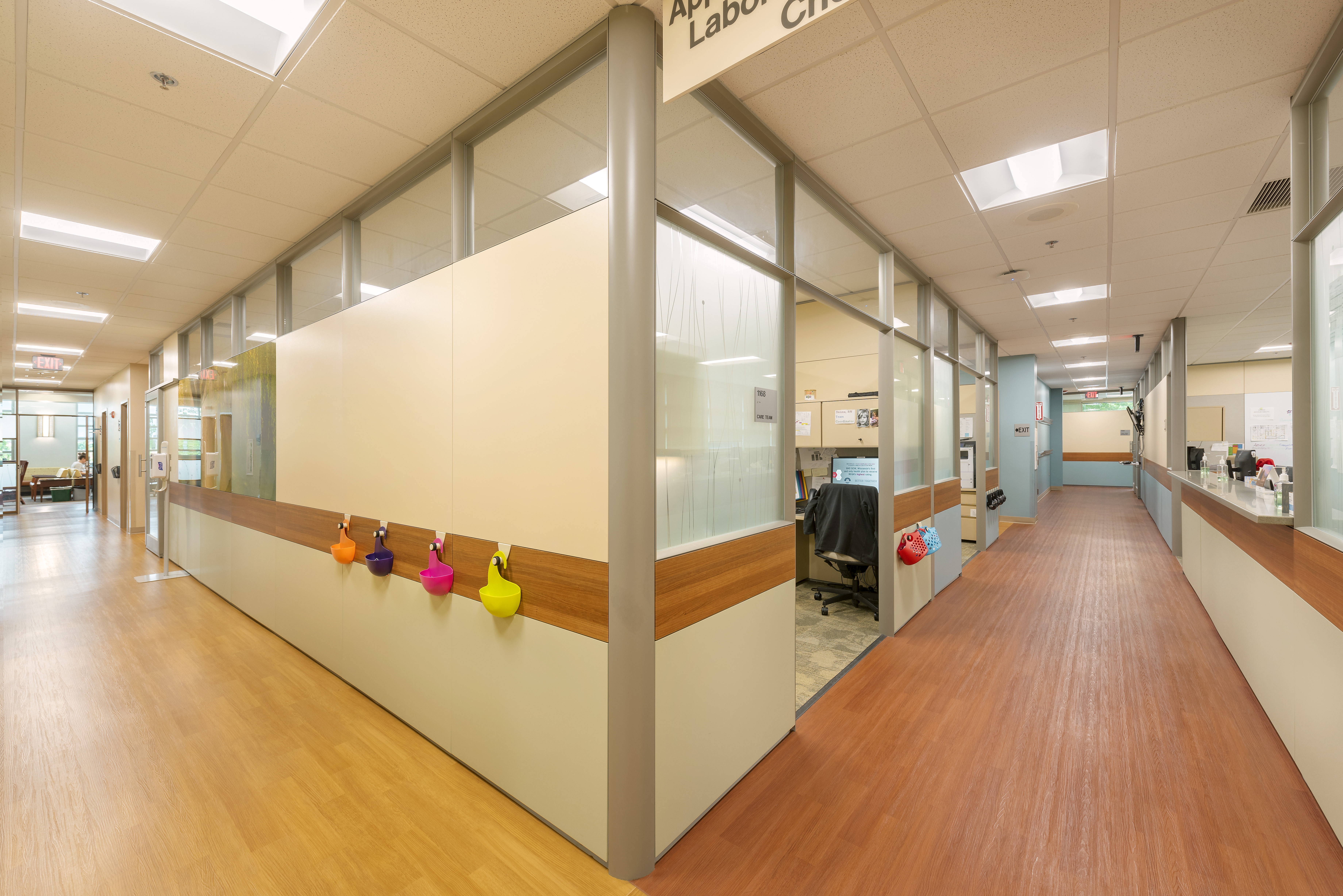 A brightly lit clinic hallway with wooden floors and glass-walled offices on the sides. Colored sanitizers are mounted on the wall outside the offices. The corridor is clean, modern, and organized, with various workstations and office supplies visible.