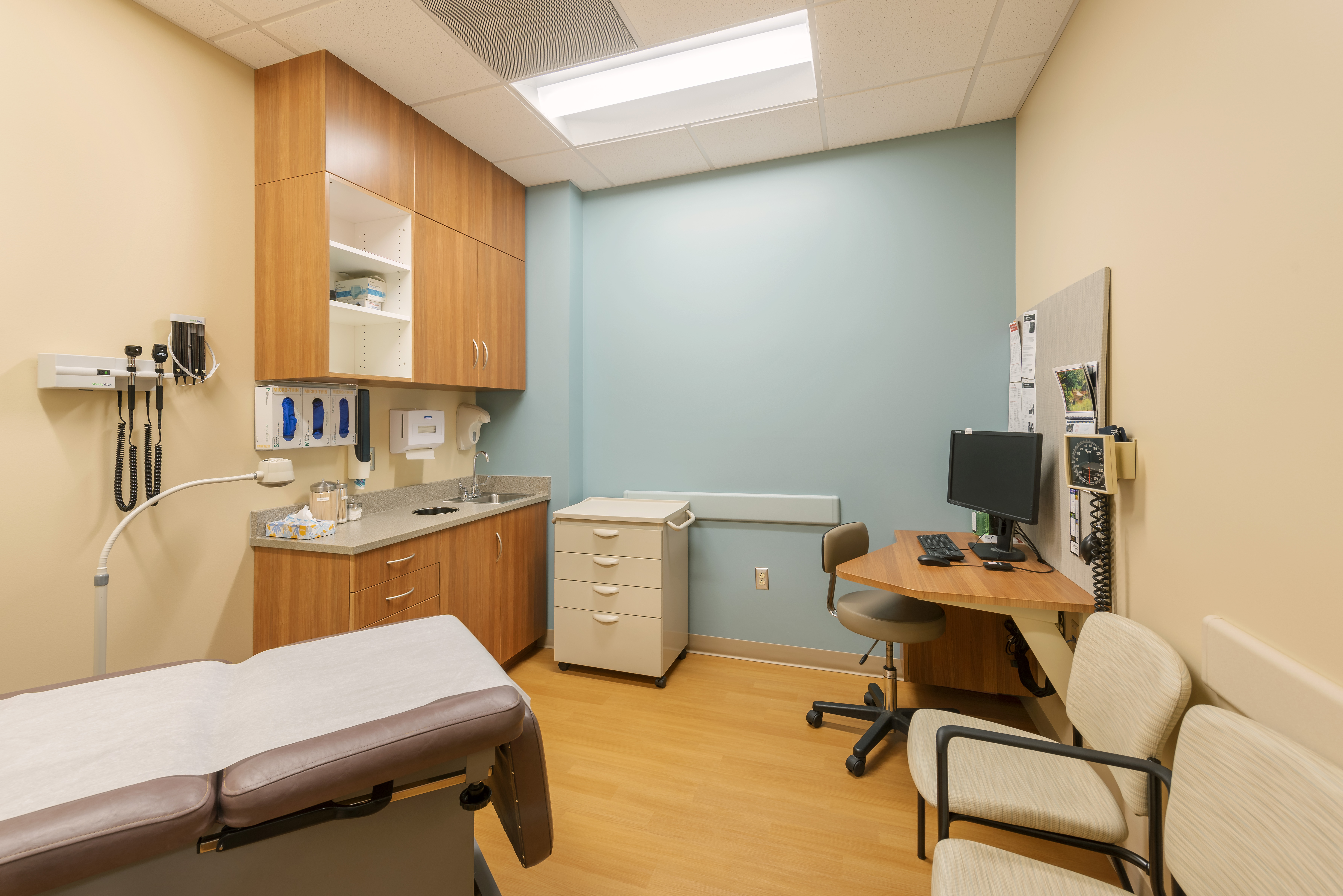 A small medical examination room with a patient bed, wooden cabinets, a beige countertop, a computer station with a chair, and medical equipment on the walls. The walls are painted light brown and teal, and the floor is wooden.