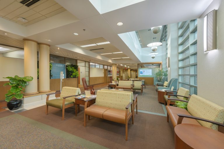 A spacious waiting area in a medical clinic with various seating options including cushioned chairs and sofas arranged around small tables. There are potted plants, large windows allowing natural light, and informational screens and board on the walls.