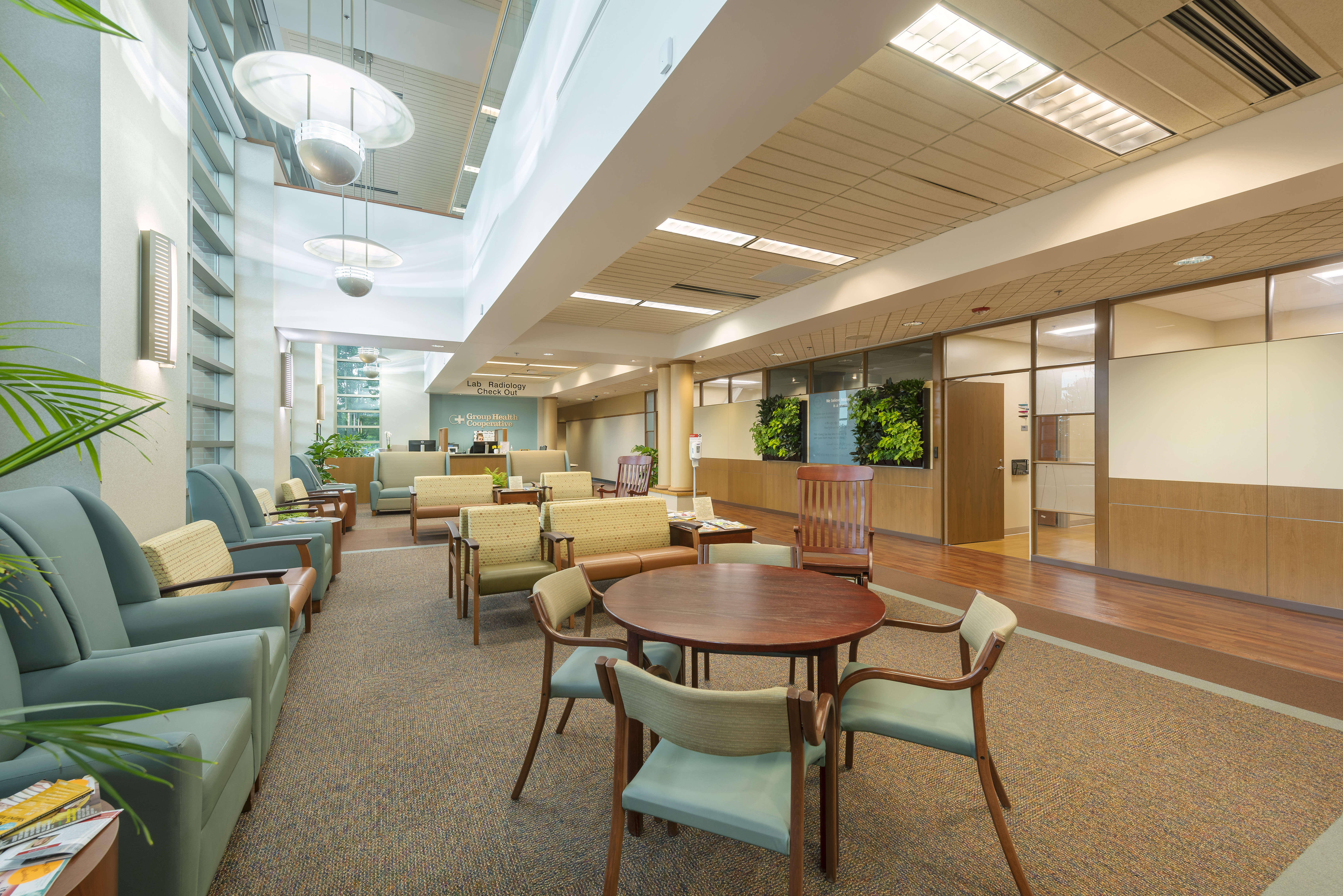 A modern waiting room with high ceilings and large windows allowing natural light. The space is furnished with green and beige armchairs, a round wooden table with chairs, and indoor plants adding a touch of nature. A reception desk is visible in the background.