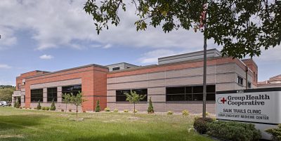 A red and grey brick building with a large sign in front reading "Group Health Cooperative Sauk Trails Clinic Complementary Medicine Center". The building is surrounded by a grassy lawn and trees, with a partly cloudy sky in the background.