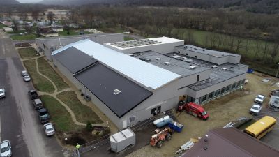 Aerial view of a large industrial building with a partially finished roof under construction. Several vehicles and construction materials are visible around the site. The surrounding area includes fields and a few houses.