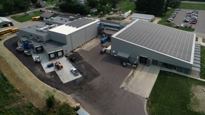 Aerial view of an industrial facility with multiple large buildings featuring solar panels on the roofs. Surrounding the facility are paved roads, grassy areas, and parking lots with parked vehicles. Trees and residential buildings are visible in the background.