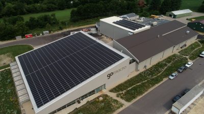 Aerial view of a modern industrial building with large solar panels on the roof. Surrounded by greenery, the site includes trucks and small vehicles parked nearby. The building is labeled with a prominent logo on the side.