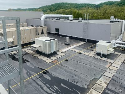 A rooftop view of a commercial building with several HVAC units and metal ductwork. The surface is flat with a few access pathways marked by pavers. A ladder leads to a higher section. Trees and hills are visible in the background.