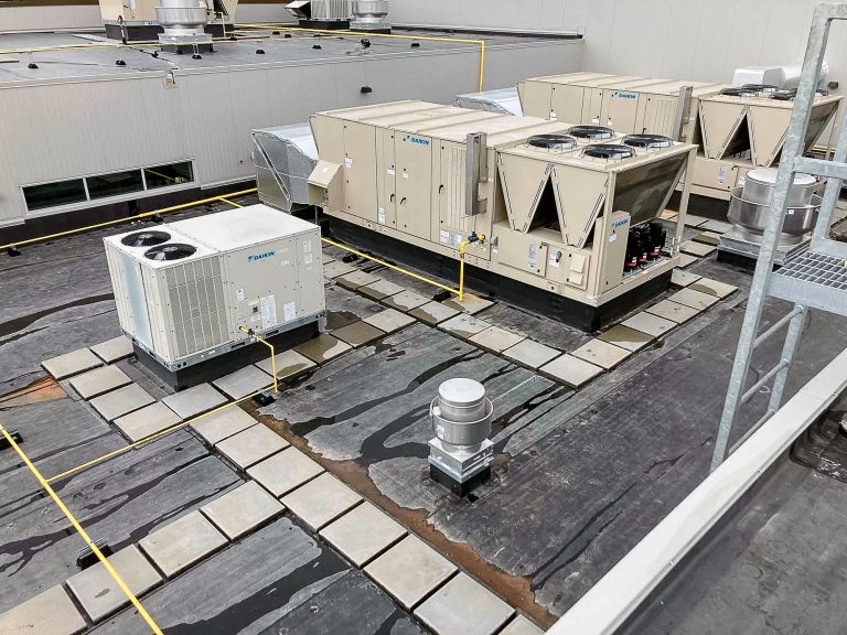 Rooftop view of a commercial building with multiple HVAC units. The equipment sits on a concrete surface with yellow markings and visible drainage areas. Adjacent walls and a metal staircase are partially visible.