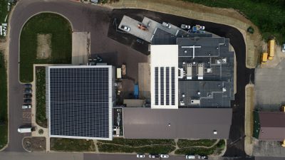 Aerial view of an industrial facility featuring a large building with solar panels on the roof. The site includes parking areas, several vehicles, and surrounding greenery.