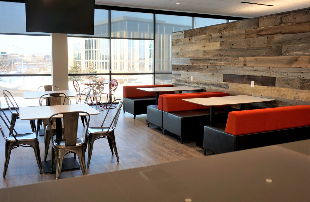 A modern cafeteria with metal chairs and square tables, alongside booth seating with red upholstery. The room features large windows allowing natural light, wooden accent walls, and a view of an exterior building. The floor is dark hardwood.