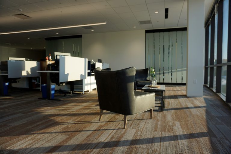 office space with two leather chairs and a small table positioned near large windows. Bright sunlight streams in, illuminating the carpeted floor. Cubicles with computers and office supplies are in the background. Frosted glass panels partially divide the area.