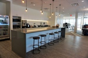 office kitchenette features a large island counter with four high chairs. Hanging pendant lights illuminate the space. The kitchenette includes a refrigerator, microwave ovens, a coffee machine, and cabinets.